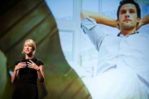 Amy Cuddy standing before a projection of a man with his hands behind his head looking powerful.